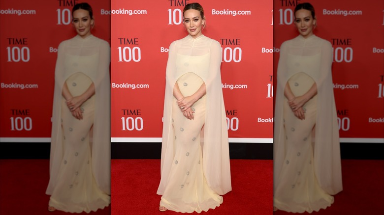 Hilary Duff posing with her hands in front of her at a red carpet event in a cream-colored gown with a cape.
