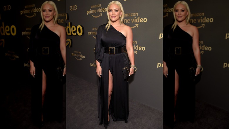 Hilary Duff smiling at a red carpet event wearing a black one-shoulder dress with a large belt.