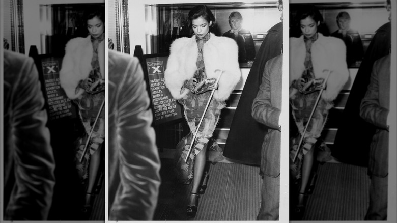 Bianca Jagger looking down wearing a white fur coat and holding a cane.