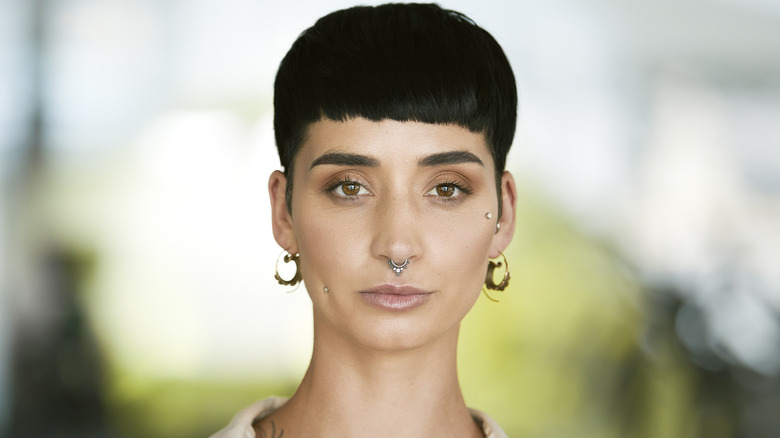A person with black blunt bangs and facial piercings posing in front of a blurred background