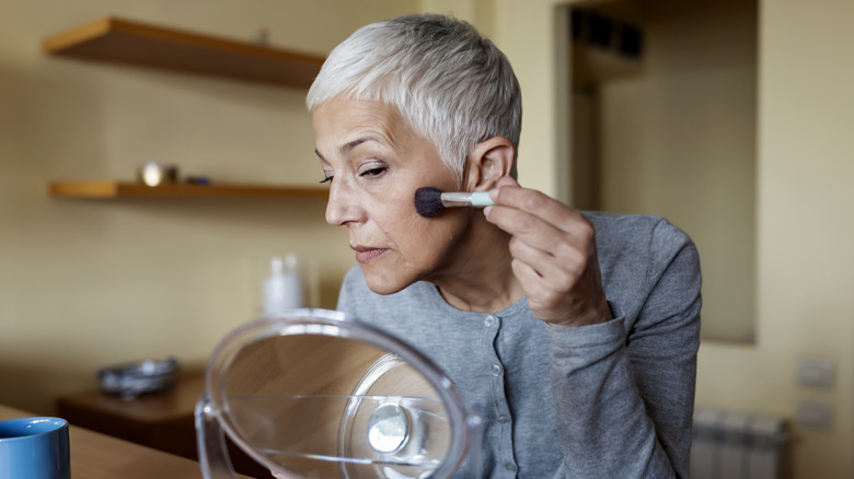 Mature woman with short hair applying blush in front of a small makeup mirror.