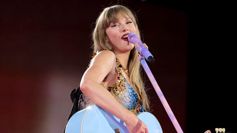 Taylor Swift wears a sparkly bodysuit as she plays the guitar and sings into a pastel microphone on stage.