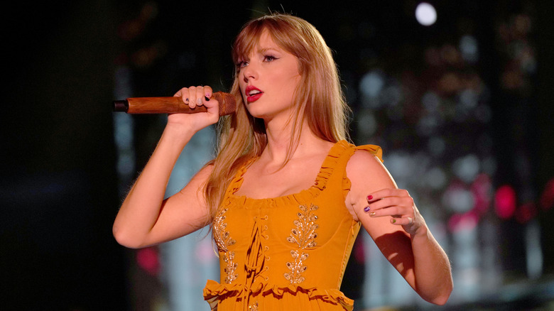 Taylor Swift wears an orange dress while singing on stage and wistfully looking out at the audience.