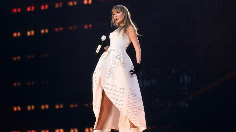Taylor Swift poses on stage while wearing a white gown and black gloves, as she holds a microphone in one hand.