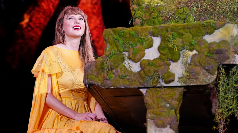 Taylor Swift smiles at the crowd while wearing a yellow dress and sitting at a moss-covered piano.