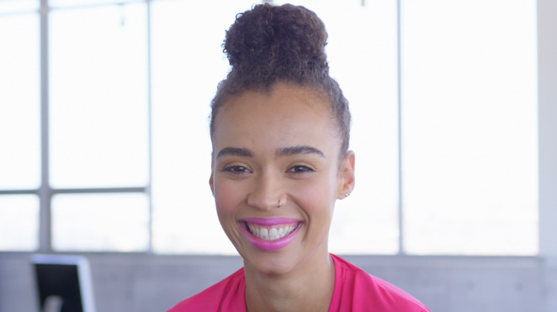 A woman with a sleek bun and pink lipstick smiling in front of windows