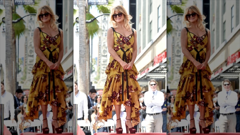 Goldie Hawn stands wearing a mustard ruffle dress and sunglasses
