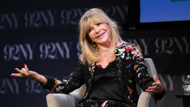 Goldie Hawn sits in a chair wearing a floral blazer and crystal choker
