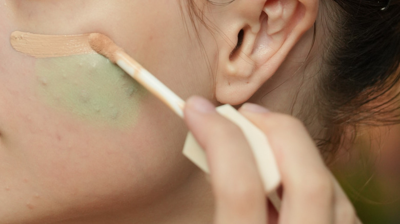 Woman applying green color corrector concealer to cheek
