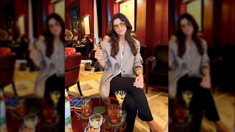 Kaitlan Collins dressed in tan blazer and pants in yellow sunglasses holding a glass of champagne