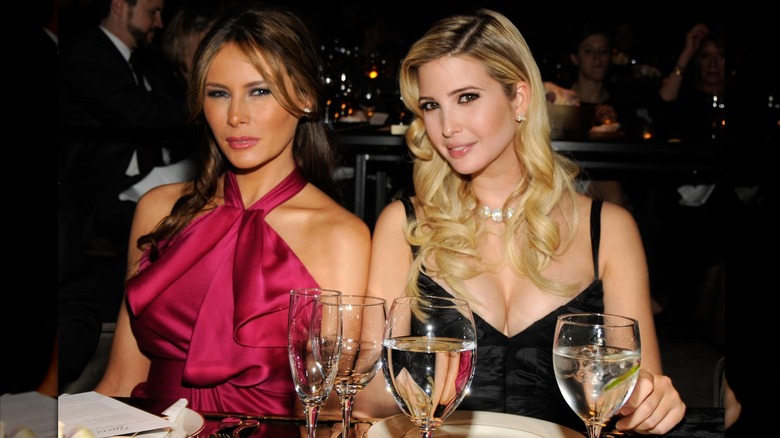 Melania and Ivanka Trump sit next to each other at a dinner table. Melania is wearing a berry colored halter-top dress with pleats. Ivanka is wearing a black dress with thin straps.