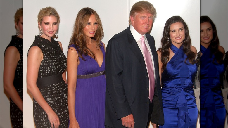 Ivanka and Melania Trump pose next to Donald Trump and Demi Moore, a fair-skinned woman with brown wavy hair. Ivanka is wearing a gray and black spotted dress with a thick belt. Melania is wearing a purple empire waist dress.