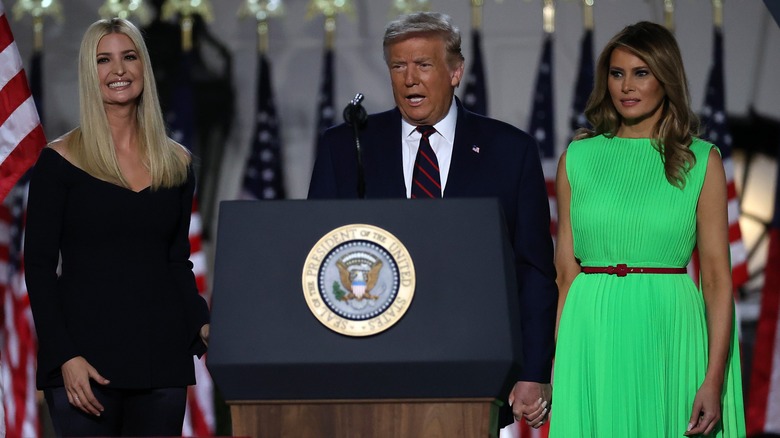 Ivanka and Melania stand next to Donald Trump, an older white man with a spray tan and light-colored hair, at a podium. Ivanka is wearing an off-the-shoulder black dress. Melania is wearing a bright green sleeveless dress.