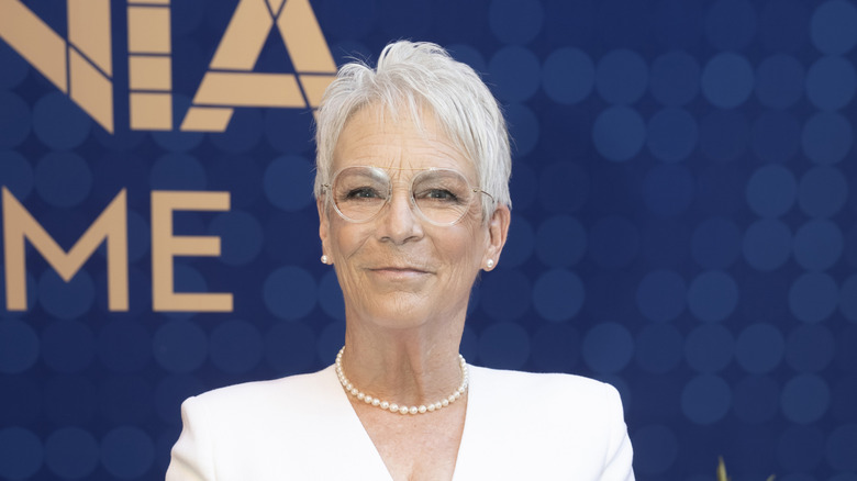 Jamie Lee Curtis posing against a blue backdrop wearing a white outfit and a pearl necklace.