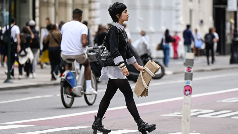 A person walks across the street in black leggings and a black sweater vest over a white blouse