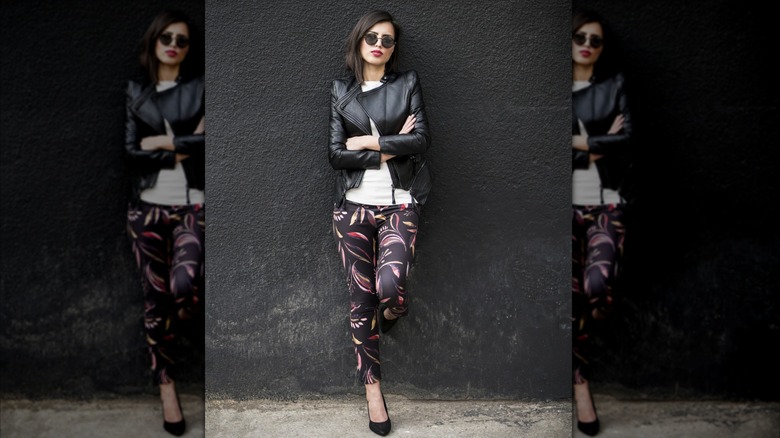 A woman stands against a wall in cool sunglasses, heels, patterned leggings and a leather jacket