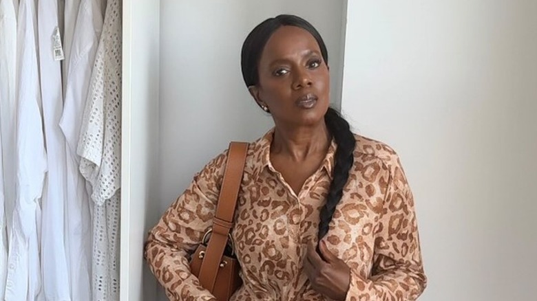 A woman with her hair in a braid posing in front of a white wall wearing an animal-print blouse.