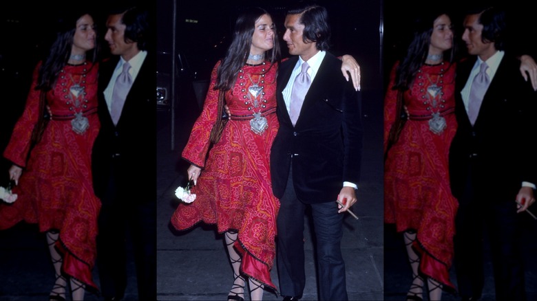 Ali MacGraw wearing a red dress, strappy sandals, and large necklace with her arm around Robert Evans wearing a suit.