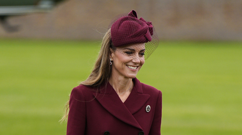 Kate Middleton smiles while walking outside wearing a brooch on her cranberry coat