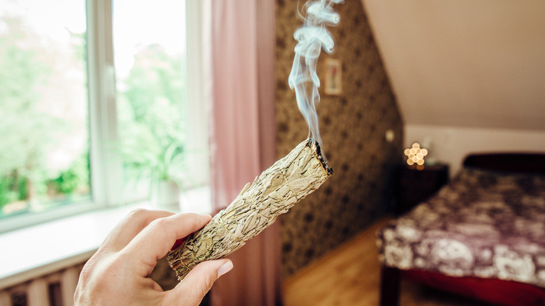 Hand holding a burning smudge stick in a bedroom.