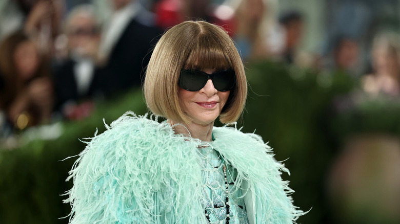 Anna Wintour smiles wearing black sunglasses and a turquoise feather jacket