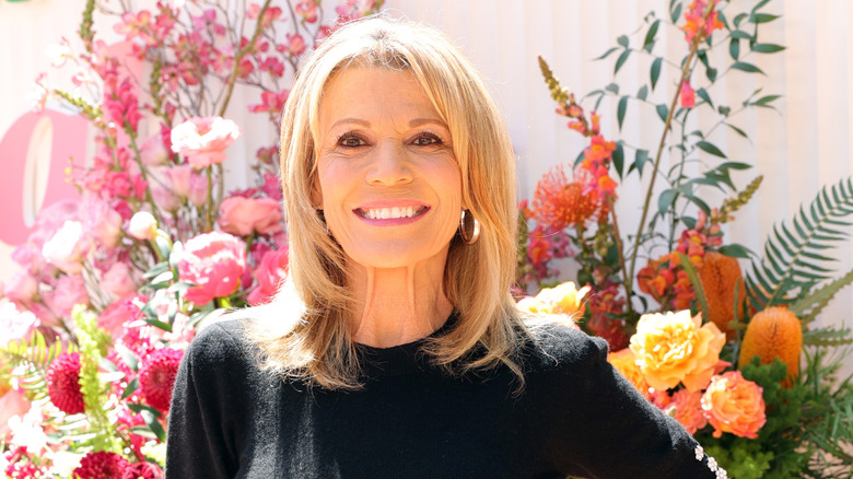 Vanna White smiling while wearing a black dress in front of a bunch of flowers.
