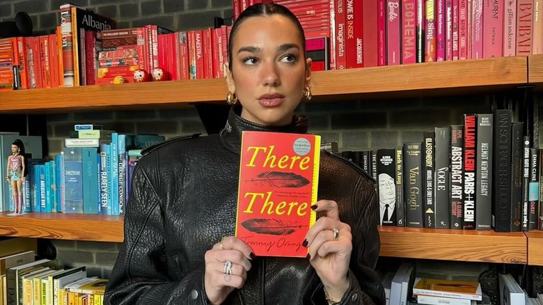 Dua Lipa holding up a book in front of a bookshelf