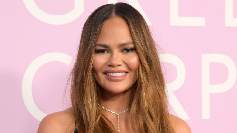 Chrissy Teigen smiles at a red carpet event wearing a diamond necklace against a pink background.