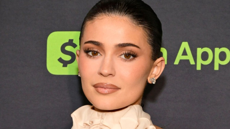 Kylie Jenner at an event. She is a young woman with fair skin, full lips, and dark hair slicked back into a bun. She is in full glam with soft brown colors.
