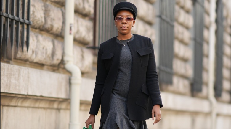 A woman walking in a dark-toned outfit, wearing a gray dress and black blazer