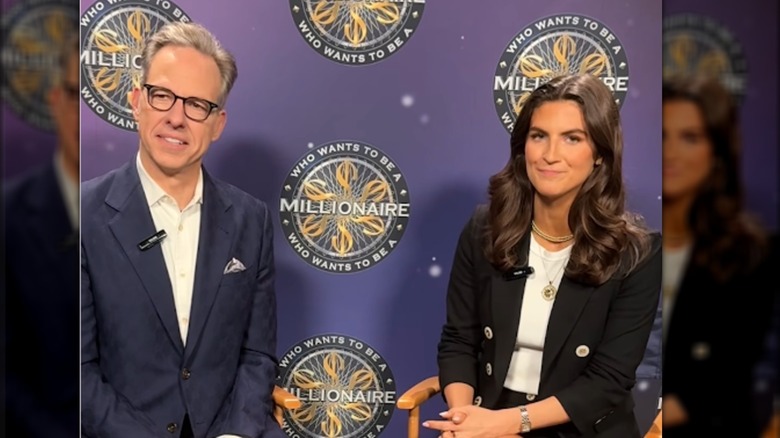Collins sits beside Jake Tapper for an interview. Tapper is an older white man with short gray hair and thick black glasses. He is wearing a navy blue suit with a white button-down. Collins is wearing a black blazer with a matching mini skirt, a white t-shirt, and a few gold necklaces. Two are made from thick gold beads and one features a large circular pendant with a sun motif on a thin gold chain.
