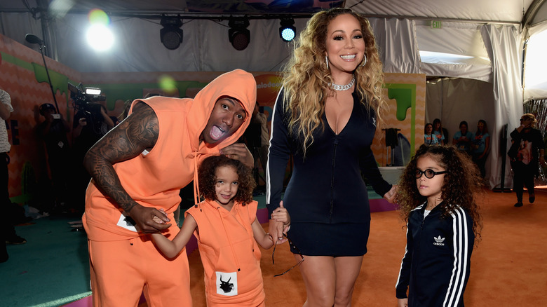 Nick Cannon wearing an orange hooded outfit and Mariah Carey in a short black dress smiling on the red carpet with children Moroccan and Monroe.