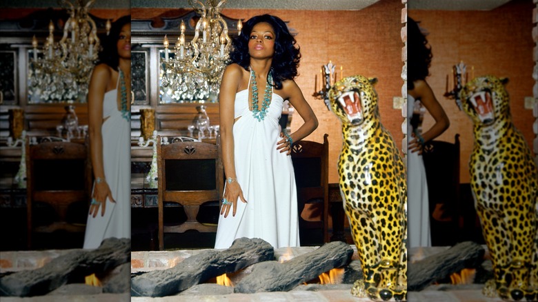 Diana Ross posing next to a cheetah statue in a flowing white dress with a green statement necklace.