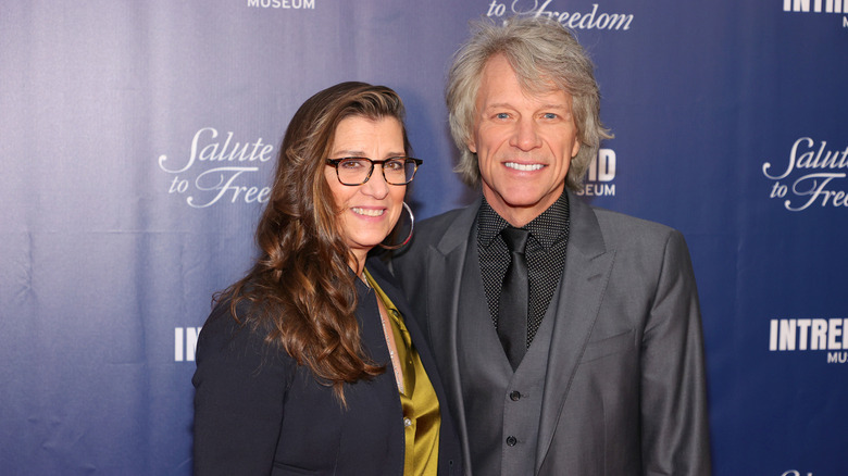 Jon Bon Jovi and his wife Dorothea Hurley posing at a red carpet event in formal outfits in front of a blue background.