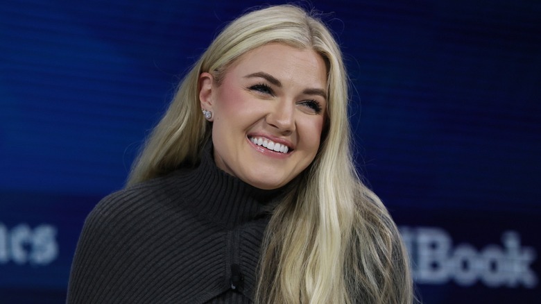 Erika Kirk sits onstage at an event. She is a woman with fair skin and long blonde hair. She is wearing a high-necked dark gray dress.
