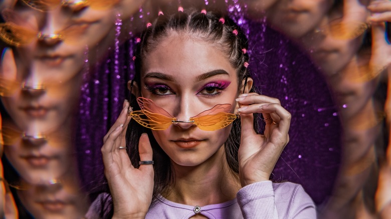 A woman looks into the camera over a pair of orange sunglasses that look like dragonfly wings. She is wearing metallic purple eyeshadow.