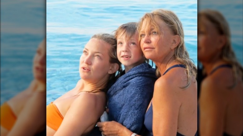 Kate Hudson, son Ryder, and Goldie Hawn pose looking upwards by the water in swimwear.