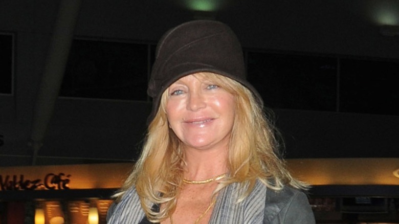 Goldie Hawn smiling wearing a black hat and gray jacket with gold necklaces.