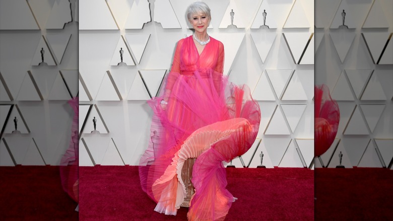 Helen Mirren posing on the red carpet in 2019 wearing a bright pink gown with a skirt that is blowing in the wind.