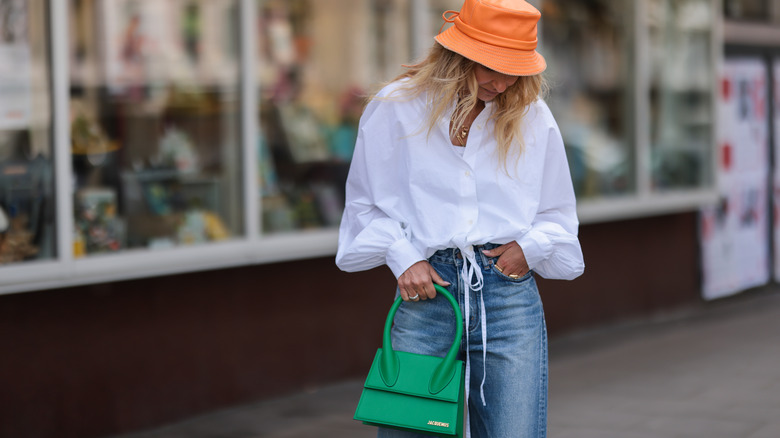 woman wearing an orange bucket hat, white button-down, blue jeans and green bag
