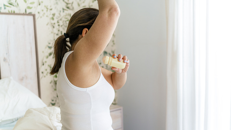 A woman applying deodorant in her underarms while sitting on a bed