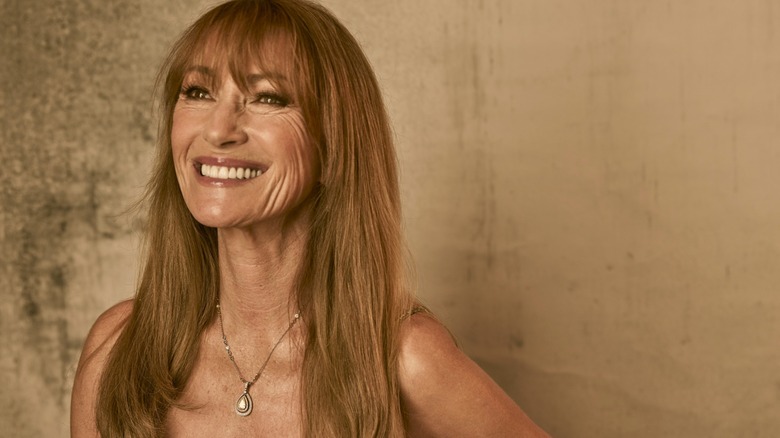 Jane Seymour smiling and wearing a pendant necklace against a brown background.