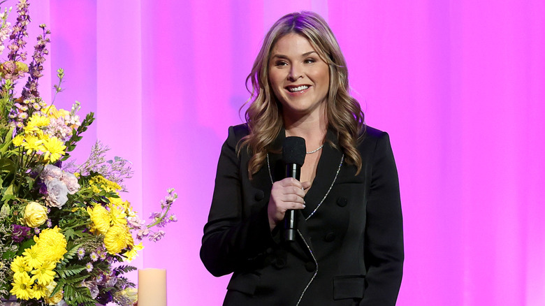 Jenna Bush Hager talking on stage while holding a microphone in front of a pink background.