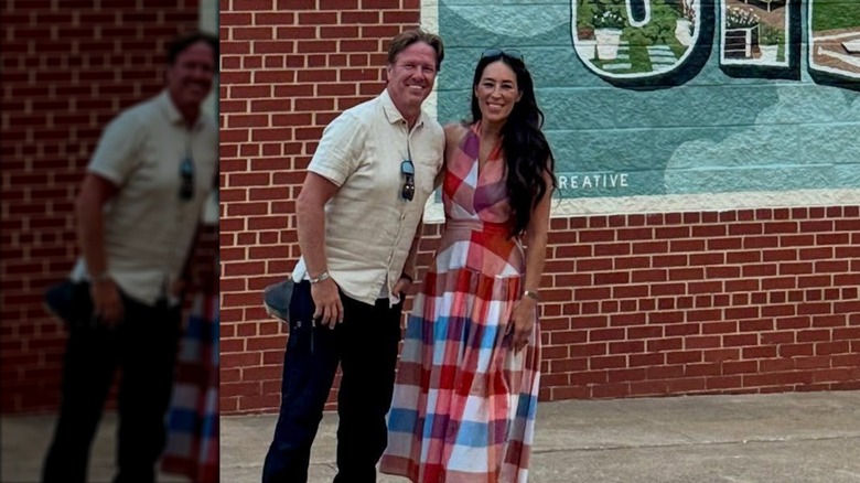 Chip Gaines in a white button-up shirt and Joanna Gaines in a plaid pattern dress posing together in front of a red brick wall.