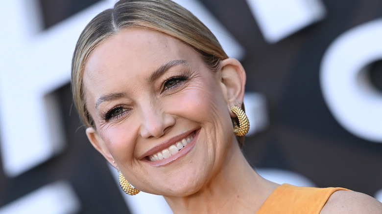 Kate Hudson smiling, wearing an orange dress with her hair in a low bun.