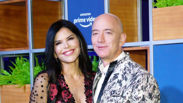 Cropped image of Lauren Sánchez Bezos and Jeff Bezos. They are standing in front of an Amazon Prime Video backdrop. Lauren is a woman with pale skin and dark hair. Jeff is a man with fair skin and a bald head.