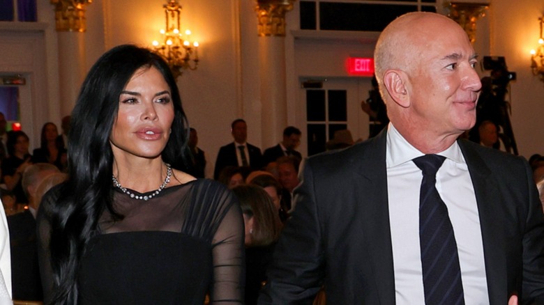Close-up shot of Lauren and Jeff Bezos at an event. Lauren is an older woman with fair skin, long black hair and full lips. This image shows the top half of her black dress. Jeff is an older man with fair skin, a bald head and no facial hair. He is wearing a plain black suit with a white button down shirt.