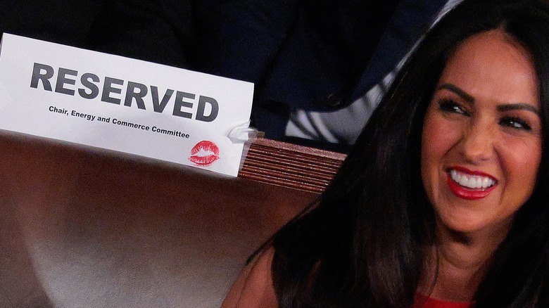 A picture of Lauren Boebert posing next to a "reserved" seat sign that she left a red kiss-mark on. She is a woman with long dark hair, a spray tan, and red lipstick.