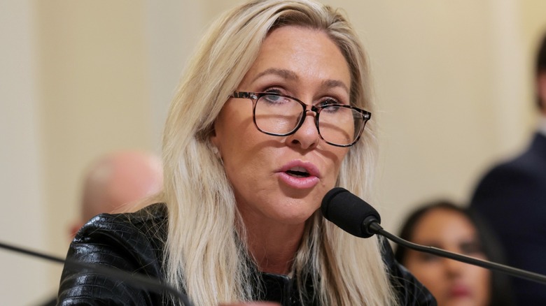 Marjorie Taylor Greene speaks at a hearing. She is an older white woman with blonde hair. In this image, she has a pair of glasses low on her nose bridge.