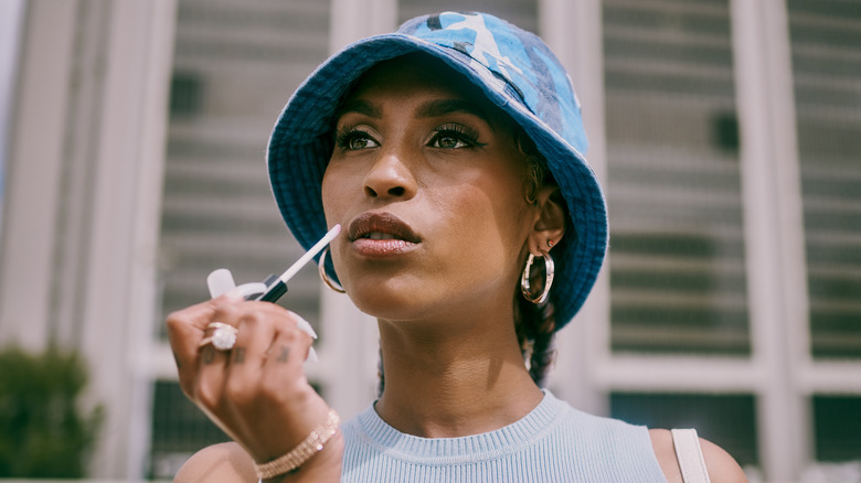 A woman applying lip gloss while wearing a blue hat